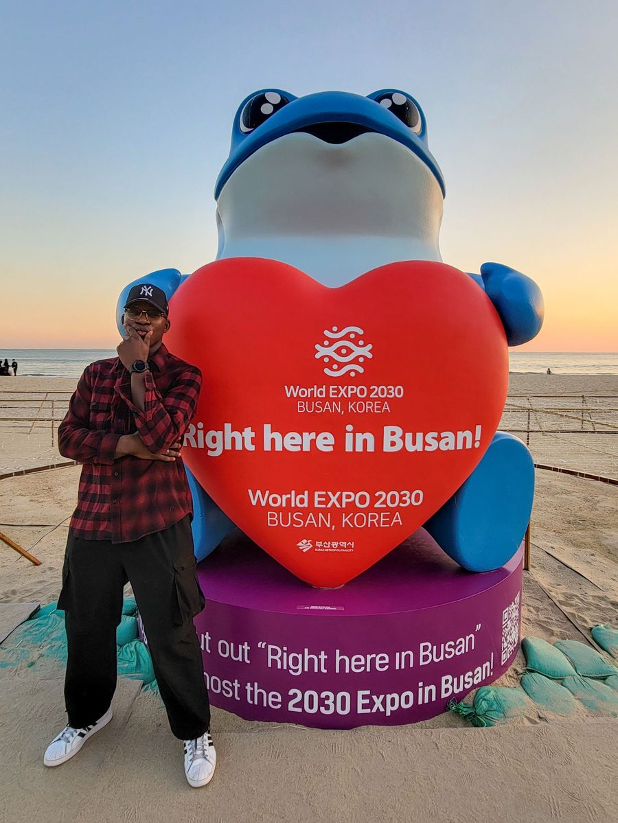 Ade at Busan beach next to a giant blue mascot holding a heart