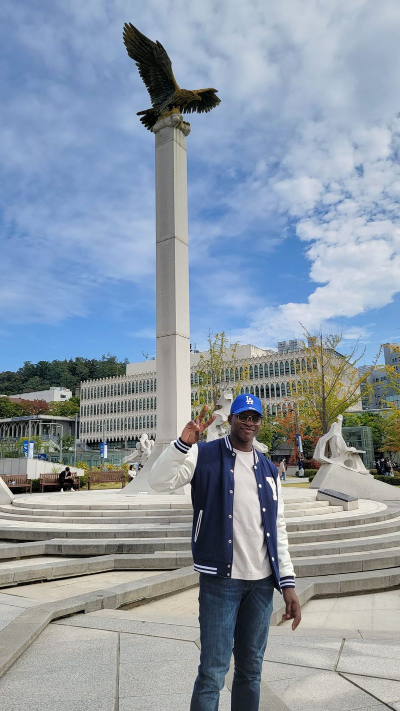 Ade in a Yonsei varsity jacket by the eagle statue, holding up a peace sign